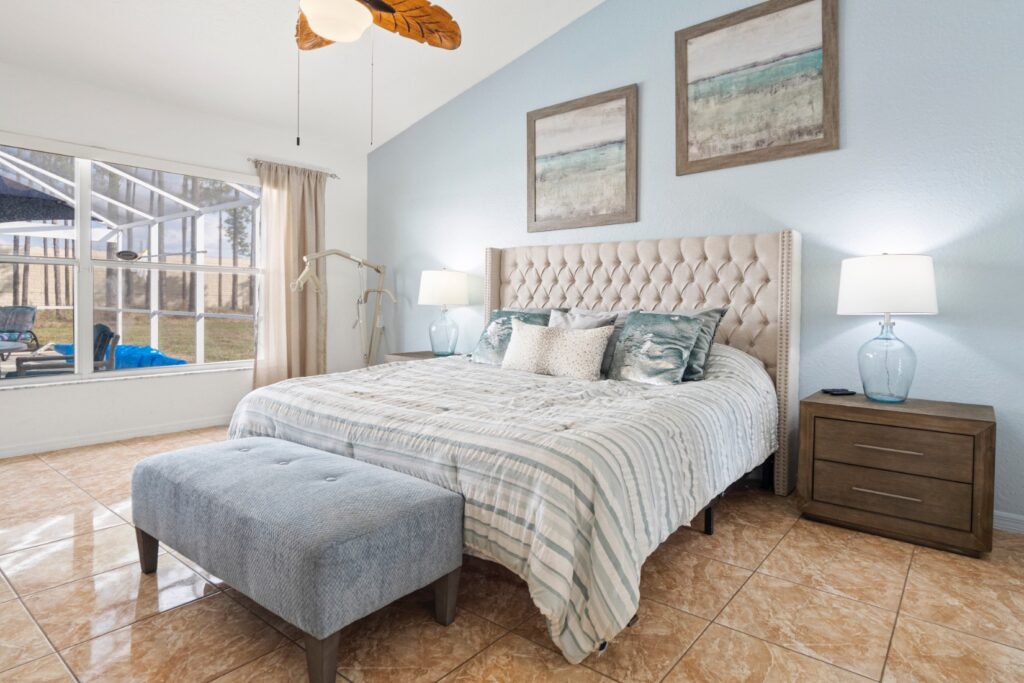 Spacious master bedroom with a tufted upholstered king bed in soft blue and cream tones, two bedside lamps, a padded bench at the foot of the bed, and a hoist positioned beside the bed available for assisted transfers. Large window overlooks the screened pool area.
