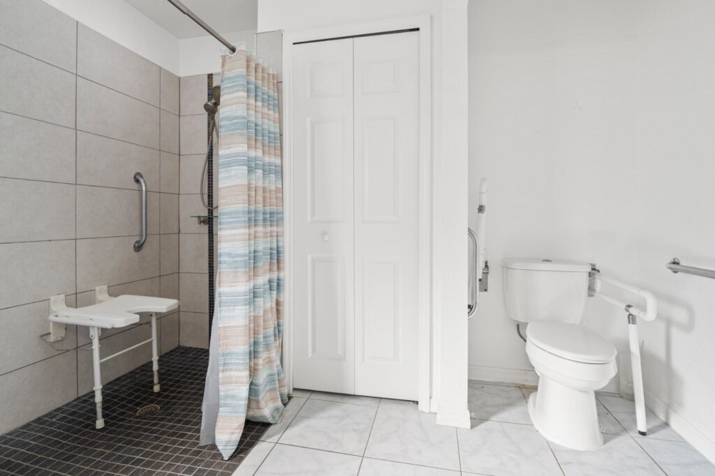 Bright white bathroom with a wooden vanity unit, large mirror, tiled floor and a shower-over-bath with a blue ocean-print curtain. A sturdy grab rail is fitted to the side of the bath for safe entry and exit.