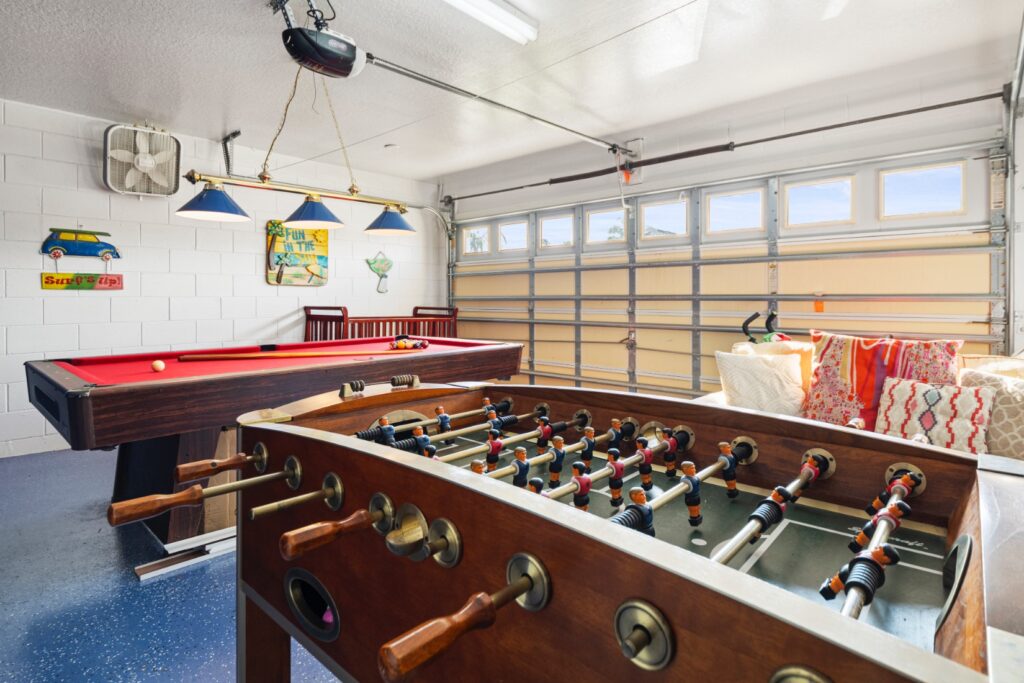 Converted garage games room with a full-size pool table, foosball table, pendant lighting, beach-themed wall decorations and a seating area with colourful cushions — ideal for family entertainment.