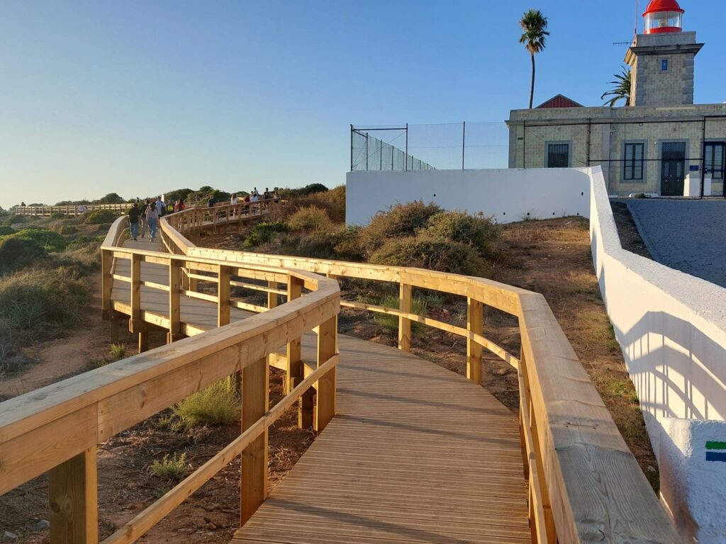 A wide, wooden boardwalk with sturdy railings curves along a coastal cliffside. To the right is a stone lighthouse building with a red lantern room and a white perimeter wall. Several people are walking along the path under a bright, clear sky.