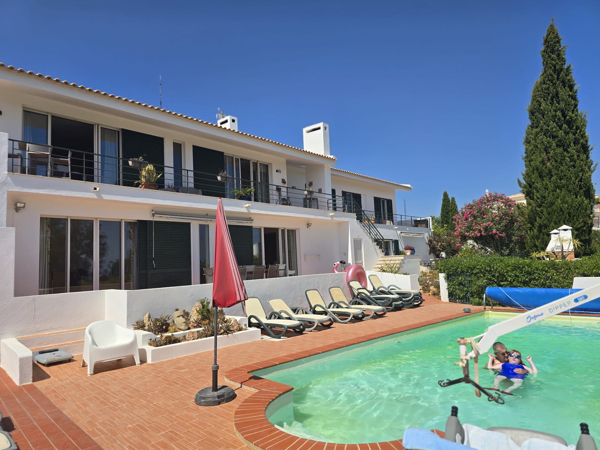 A bright, modern guesthouse with a large outdoor swimming pool in front. The pool includes a mechanical pool lift assisting a person into the water, highlighting accessible swimming सुविधities. Surrounding the pool are multiple sun loungers, umbrellas, and spacious, level tiled areas, with the accommodation building featuring balconies and large sliding doors overlooking the pool.