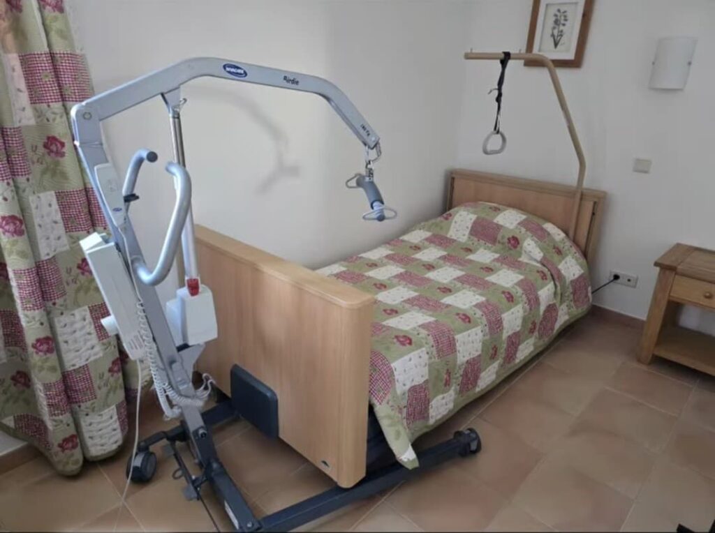 A private room featuring an adjustable wooden-frame profiling bed with a patterned green and red quilt. Positioned at the foot of the bed is a silver "Birdie" mobile floor hoist with a spreader bar, used for patient transfers.