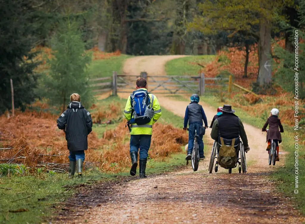 A group of people, including a man using a manual wheelchair and several children on bicycles, enjoying an accessible wide dirt trail in a forest setting during autumn.
