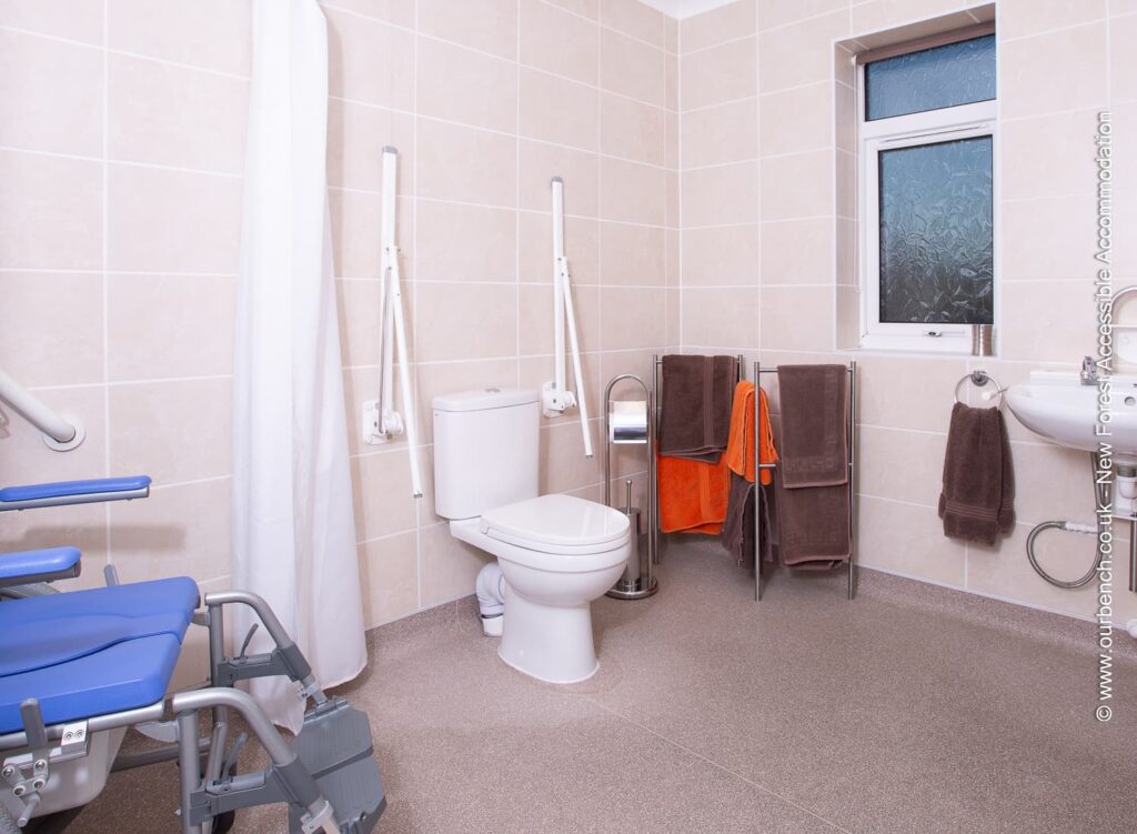 Accessible bathroom with a roll-in shower, toilet equipped with drop-down grab rails on both sides, and a blue shower chair. Includes a wall-mounted washbasin and non-slip flooring.