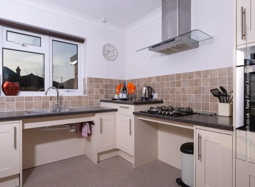 Wheelchair-accessible kitchen with lowered countertops and open knee space under the sink and stovetop for easy access. Features include a modern extractor hood and cream-colored cabinetry.