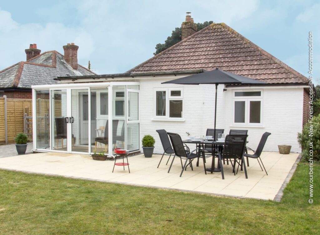 A flat, paved backyard patio with a black outdoor dining set and umbrella, next to a white bungalow with a glass-enclosed sunroom.