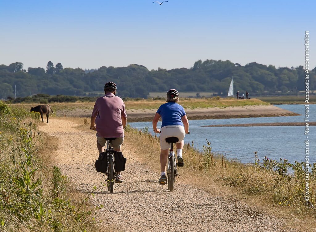 Two people cycling on a flat, gravel coastal path next to the water on a sunny day. A sailboat and a calm shoreline are visible in the background, illustrating accessible level cycling routes.