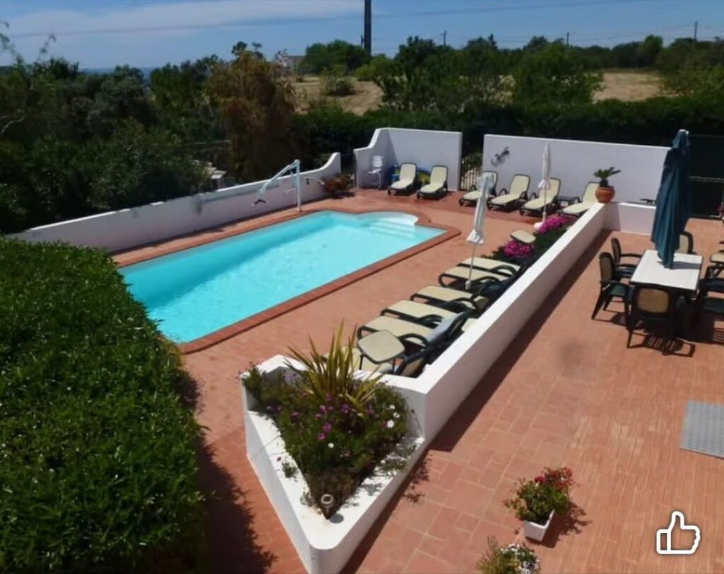 An outdoor swimming pool area with terracotta tiled flooring and white walls, surrounded by greenery. Sun loungers are neatly arranged along the poolside, and a handrail provides supported entry into the water. The space appears level and step-free, with wide pathways and open seating areas designed for accessible movement.