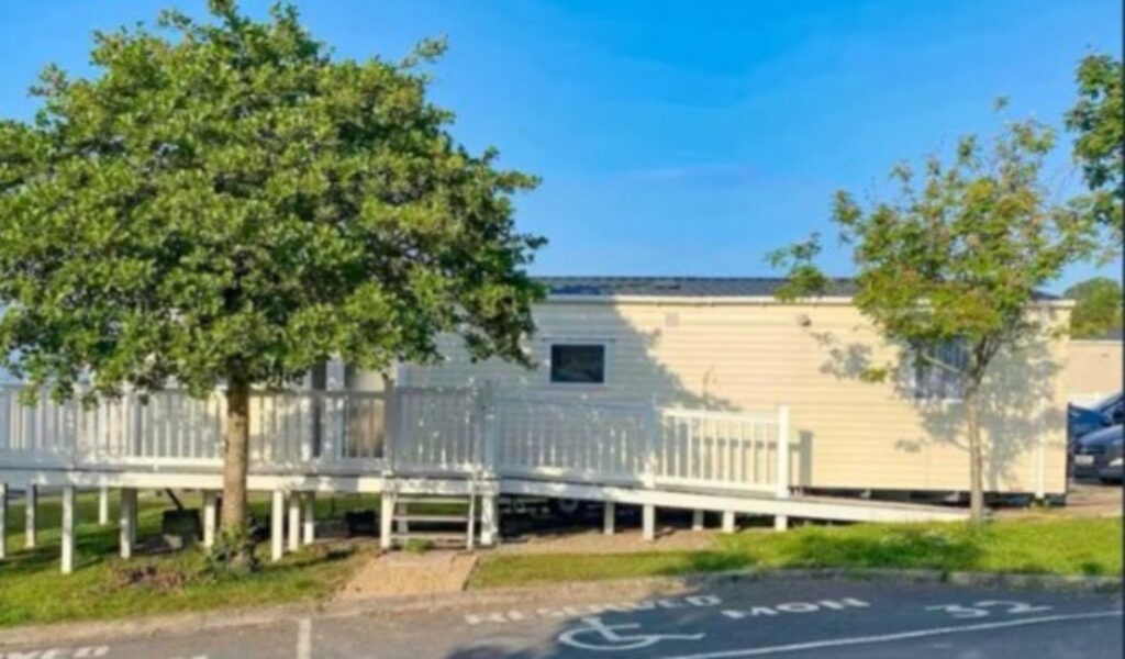 Exterior view of the fully adapted caravan on a holiday park, showing a wide accessible ramp leading up to the decked veranda, surrounded by trees, and a reserved accessible parking bay directly in front.