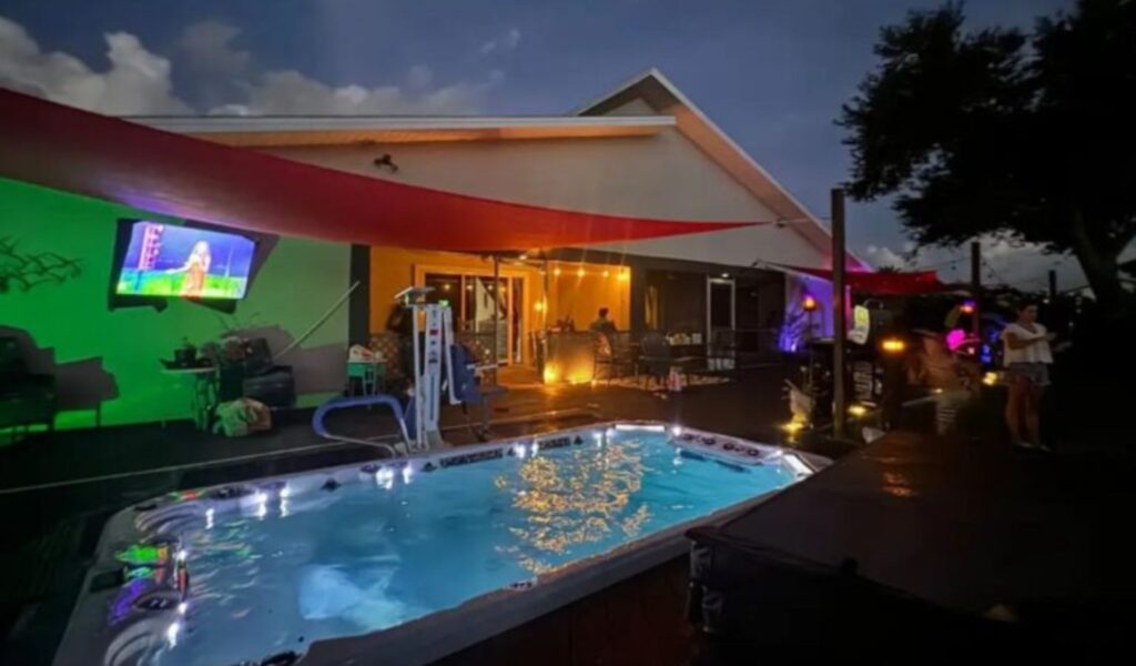 Evening view of an accessible villa's backyard in Cape Coral, featuring a well-lit swim spa with a pool lift and a mounted outdoor TV. The patio area has comfortable seating, string lights, and red sun shades, creating a cozy and inviting atmosphere.