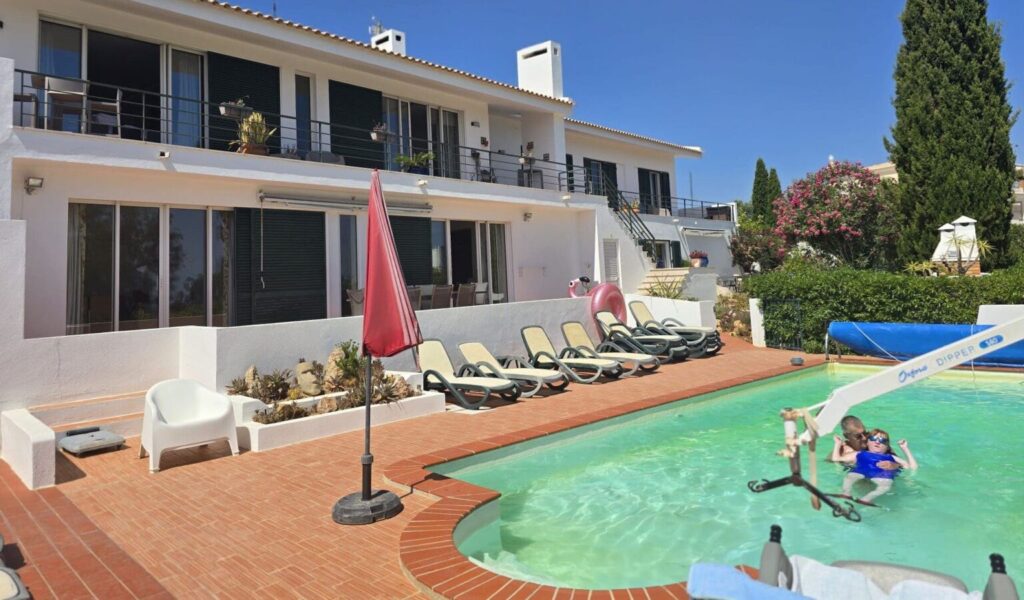 A sunny outdoor pool area in front of a white two-story villa with balconies and sliding glass doors. Several lounge chairs line the poolside on a tiled patio, along with a red umbrella and scattered outdoor seating. In the pool, a person assists a child using a pool lift device. The scene is surrounded by greenery and flowering plants under a clear blue sky.