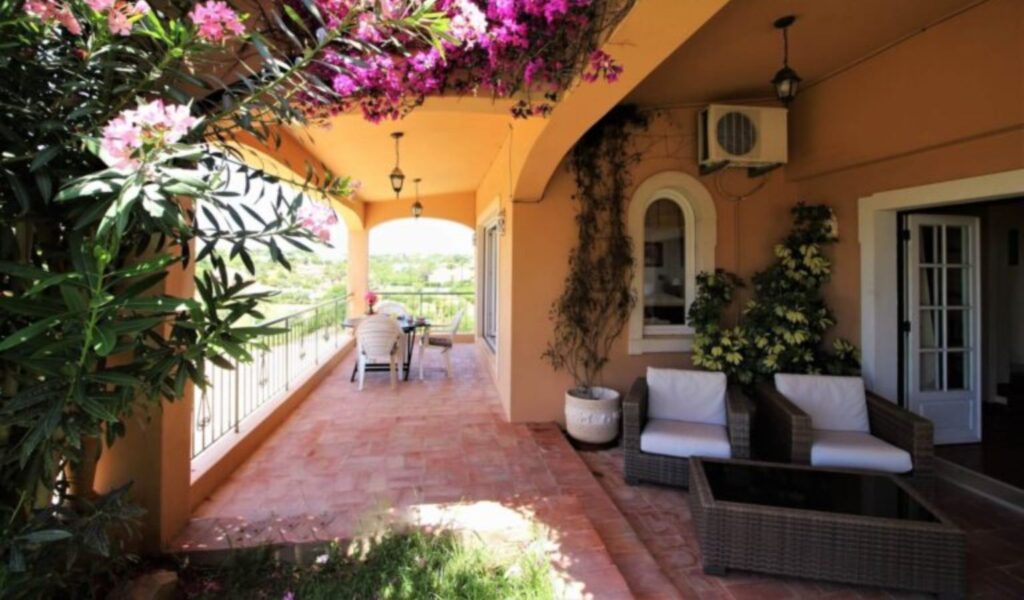 A warm-toned covered terrace with terracotta flooring and arched openings, decorated with lush greenery and vibrant pink flowering vines overhead. The space includes a small dining table and chairs near a railing with a scenic view, as well as cushioned outdoor seating with a coffee table. Potted plants and wall-mounted lighting add to the cozy, Mediterranean-style atmosphere.