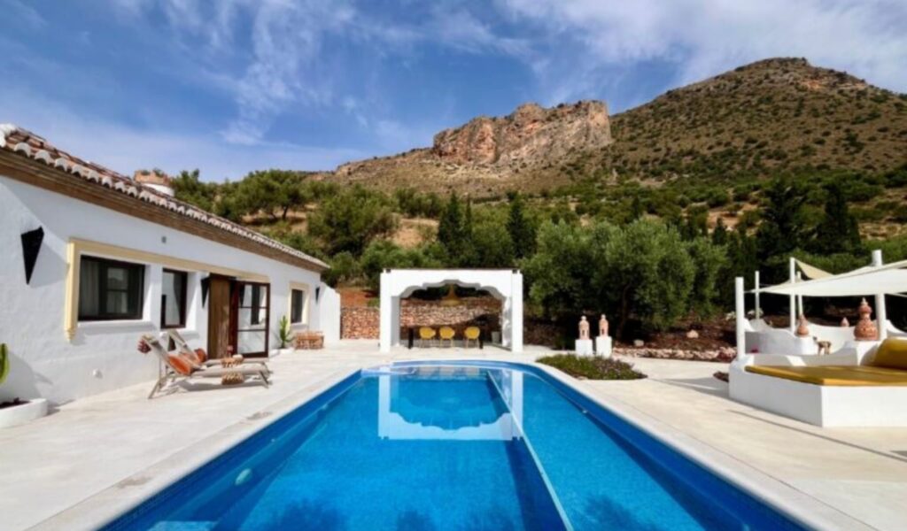 A luxurious white Mediterranean-style villa with a long rectangular lap pool in the foreground, surrounded by a light stone terrace. To the left, lounge chairs sit beside the whitewashed building with terracotta roof tiles and wooden doors. In the center background, a rustic arched pergola with a stone bar and yellow stools is framed by olive trees. To the right, a daybed with a cream umbrella and mustard cushions rests near decorative ceramic urns. A dramatic rocky hillside with sparse green vegetation rises under a partly cloudy blue sky.
