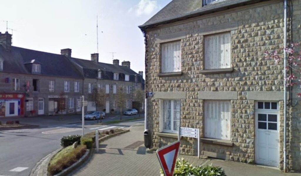 Quiet street corner in Sourdeval, Normandy, showing the traditional stone-built property on a peaceful town square with nearby shops visible in the background.