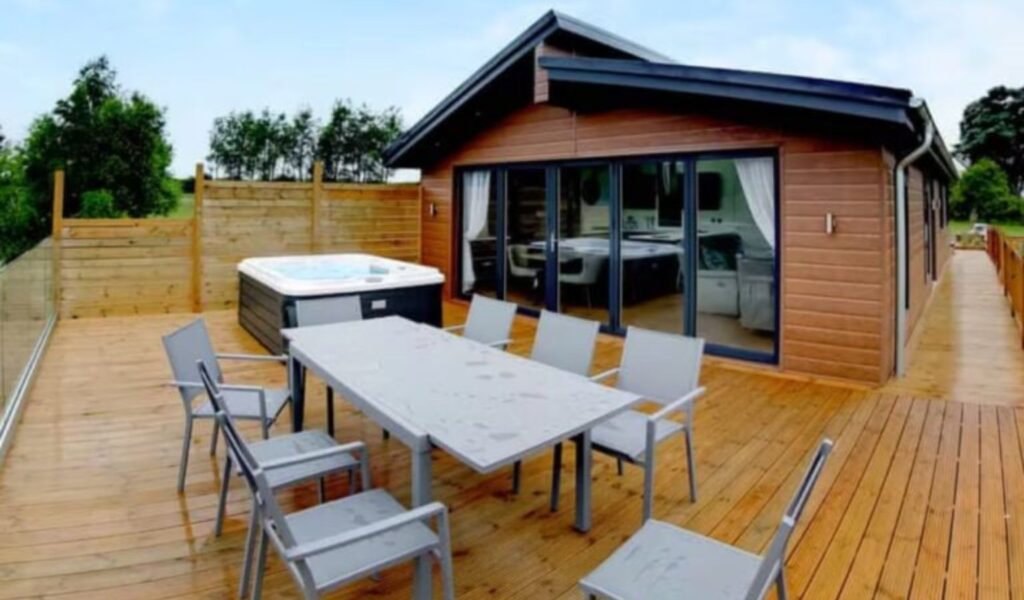A modern wheelchair-friendly cabin at Noah's Retreat in Lincolnshire featuring a spacious wooden deck with outdoor seating for six and a private hot tub. This accessible holiday home offers a relaxing retreat surrounded by greenery.