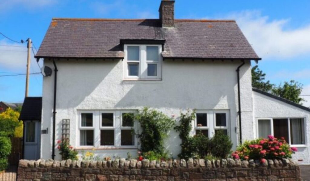A small white cottage-style house with a steep gray roof and central chimney, featuring multiple windows, a side extension, and a front garden with flowering plants behind a low stone wall under a clear blue sky.