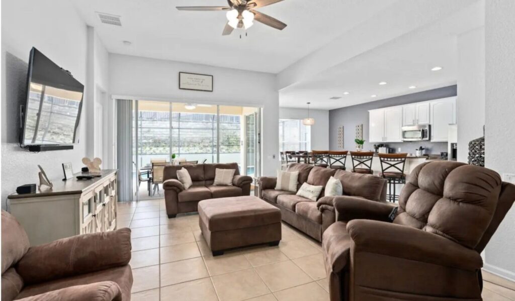 A bright, open-concept living room with beige tile floors and light gray walls, featuring a brown sofa set with matching armchairs and ottoman arranged around a wall-mounted TV. Sliding glass doors at the back lead to a patio with outdoor seating and a view of a hillside. The space flows into a modern kitchen with white cabinets, a breakfast bar with high chairs, and recessed lighting.