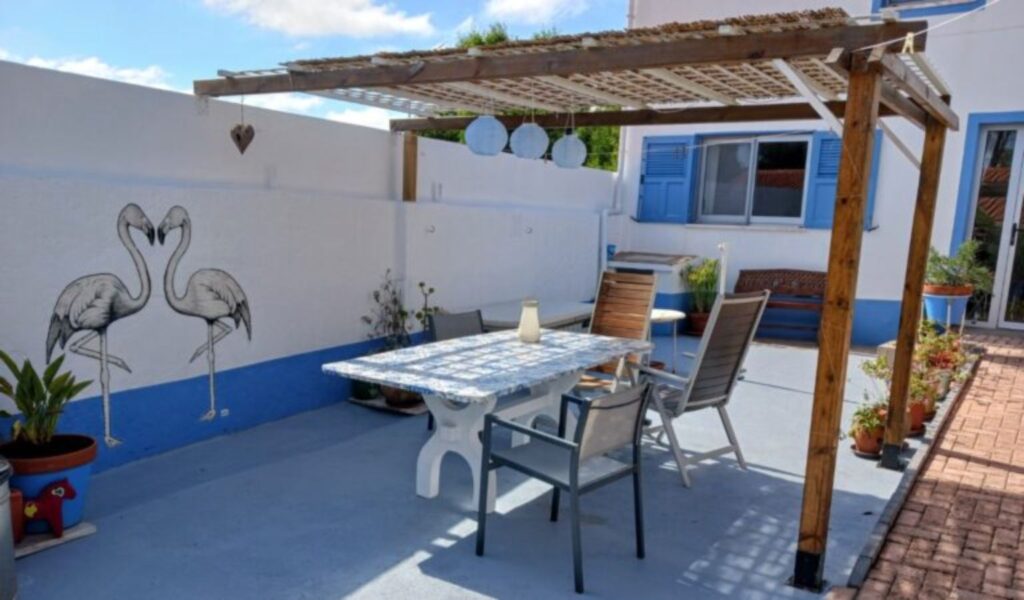 A bright outdoor patio with a wooden pergola providing partial shade over a table and chairs. The space has white walls with blue trim and features a painted mural of two flamingos facing each other. There are potted plants along the edges, a small seating bench near a window with blue shutters, and hanging lantern-style lights under the pergola. The setting appears sunny and relaxed, with a clean, coastal-style design.