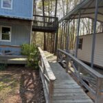 View of a long wooden accessibility ramp with handrails leading from the driveway to the elevated side porch of a two-story blue cabin.
