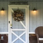 Close-up of the cabin's white front door with a decorative wreath, featuring a 'Cabin Sweet Cabin' sign, porch lights, and a keyless entry smart lock.