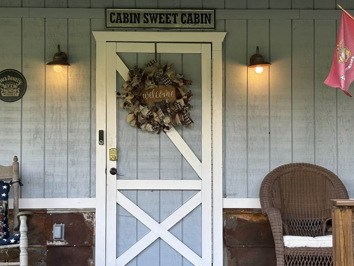 Close-up of the cabin's white front door with a decorative wreath, featuring a 'Cabin Sweet Cabin' sign, porch lights, and a keyless entry smart lock.