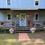 Full front view of the light blue two-story cabin featuring a wide covered porch with rocking chairs and a small set of wooden steps leading to the lawn.