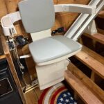 A close-up view of a motorized chair lift with a grey seat and armrests, parked at the base of a wooden staircase over a star-patterned rug.
