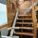Looking up a set of open-tread wooden stairs equipped with a motorized chair lift rail. The walls are decorated with rustic signs and a black-and-white checkered curtain.