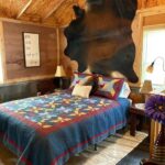 The second-floor loft bedroom featuring a bed with a colorful patchwork quilt, a cowhide rug mounted on the wall as a headboard, and rustic wood-paneled walls.