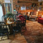 Interior view of the rustic living area with wood walls, featuring a dining table with four black chairs, a red sofa, and a large patterned rug.