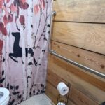 Close-up of a bathroom wall with wood paneling, featuring a long horizontal stainless steel grab bar and a vertical grab bar next to a shower curtain with a deer and forest motif.