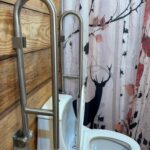 A white toilet equipped with dual foldable stainless steel safety rails on both sides, mounted against a rustic wood-plank wall.