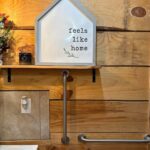 Close-up of a rustic wood-plank wall featuring two stainless steel grab bars (one vertical, one horizontal) beneath a small shelf with a 'Feels Like Home' sign.