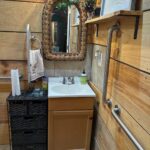 A compact bathroom vanity with a white sink and wood cabinet, surrounded by multiple stainless steel grab bars on the adjacent walls and a wicker-framed mirror above.