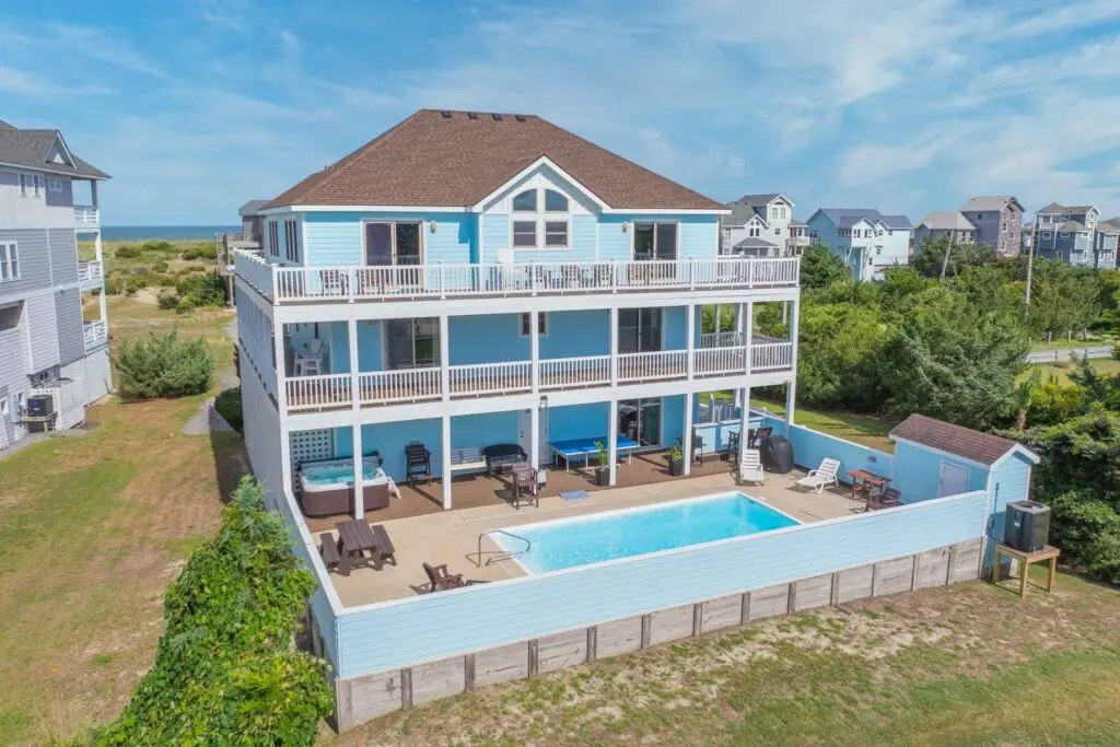 Large light blue three-story beach house with an accessible pool area. The fenced backyard features a swimming pool with a metal grab rail, a hot tub, and a level patio with seating, with the ocean visible in the distance.
