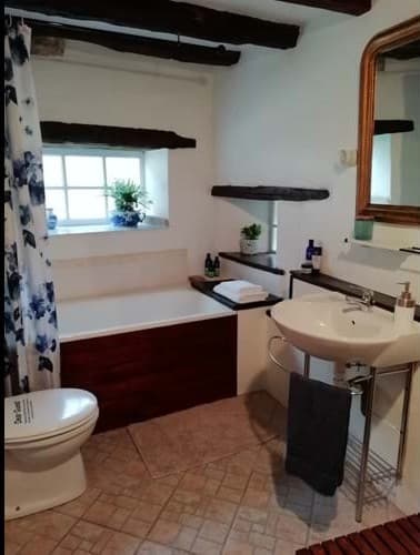 A secondary bathroom with a rustic feel, featuring exposed dark wood ceiling beams and a tiled floor. It includes a built-in bathtub with a dark wood panel, a classic white pedestal sink, and a toilet. The room is decorated with small plants and a floral shower curtain.