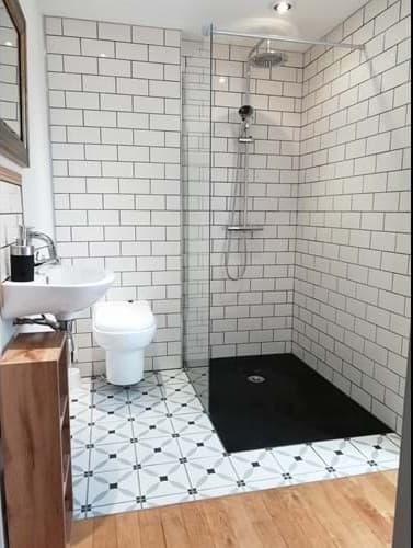Modern accessible bathroom featuring a large walk-in wet room shower with a black non-slip floor and white subway wall tiles. The transition from the wooden floor to the tiled bathroom is level, and the toilet and sink are positioned with clear floor space.