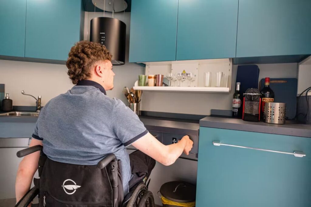A Wheelchair user in an accessible kitchen with teal cabinets, lowered countertops, and open floor space allowing wheelchair maneuverability