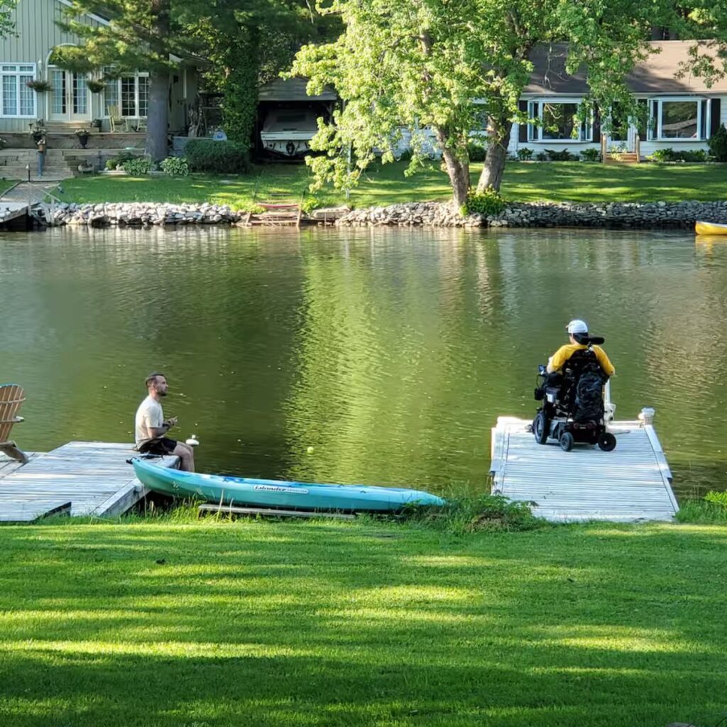 A lush green lawn leading down to a calm river. Two wooden docks extend into the water. On the left dock, a person sits near a blue kayak; on the right dock, a person in a power wheelchair is positioned safely at the end of the pier.