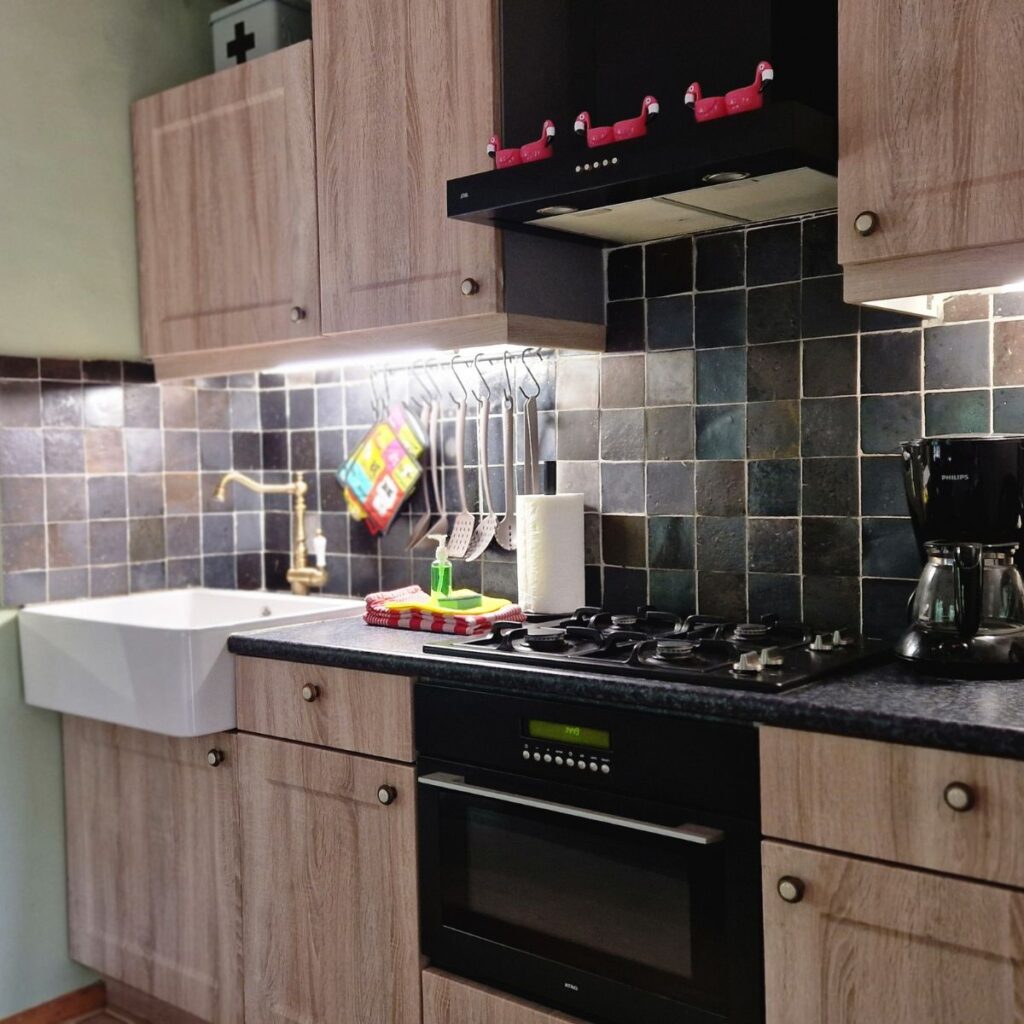 A galley-style kitchen with light wood cabinetry and dark tile backsplash. The oven and stovetop are positioned at counter height for easier access. A deep farmhouse sink is located next to the cooking area, and kitchen utensils are hung on an easy-reach rail beneath the upper cabinets.