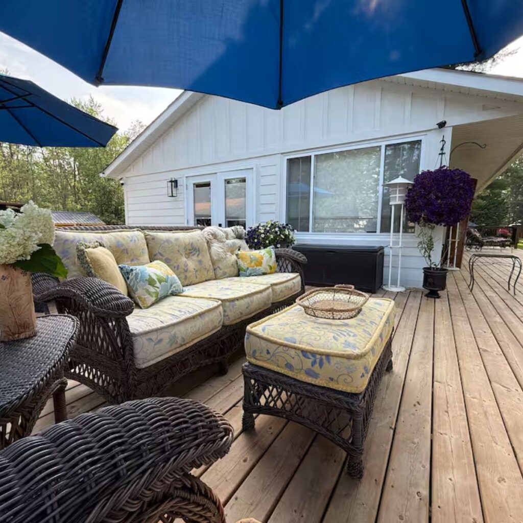 A spacious wooden deck with wicker patio furniture and yellow floral cushions. A large blue patio umbrella provides shade. The deck is wide and flat, offering plenty of turning space for a wheelchair, and is bordered by a dark safety railing.