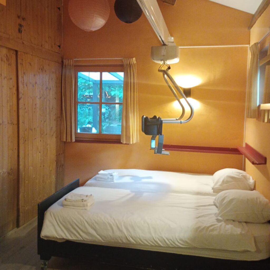 A bedroom featuring two twin beds positioned side-by-side. A ceiling-mounted track hoist system is installed over the beds to assist with transfers. The room has warm orange walls, a large window, and wide floor space at the foot of the beds.