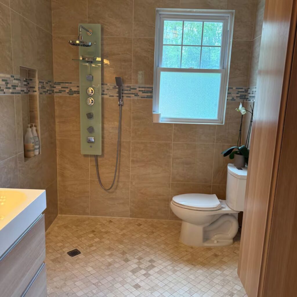 A modern bathroom featuring a zero-entry (curbless) walk-in shower with beige tiling. The shower includes a handheld showerhead. The open floor plan allows for a seamless transition between the vanity, toilet, and shower area.