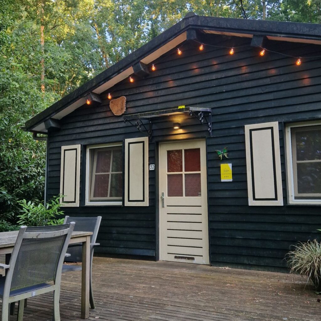 A dark wood cottage with a wide, flat wooden deck at ground level. The front door has a low threshold and is flanked by large windows with cream-colored shutters. A string of warm lights hangs from the eaves over an outdoor dining table.