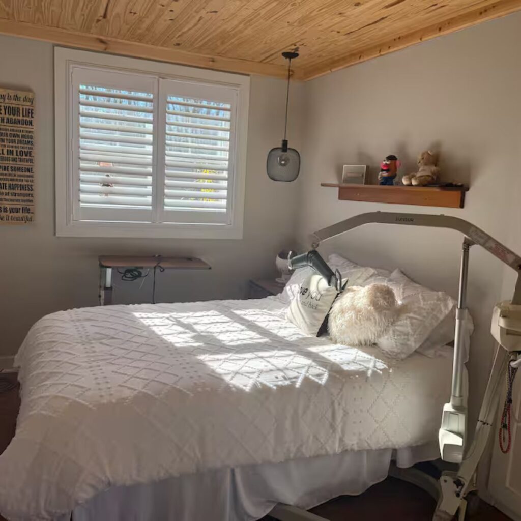 A well-lit bedroom featuring a white quilted bedspread and a wood-paneled ceiling. An overhead patient lift is positioned next to the bed. To the left, a height-adjustable rolling bedside table sits under a window with white shutters.