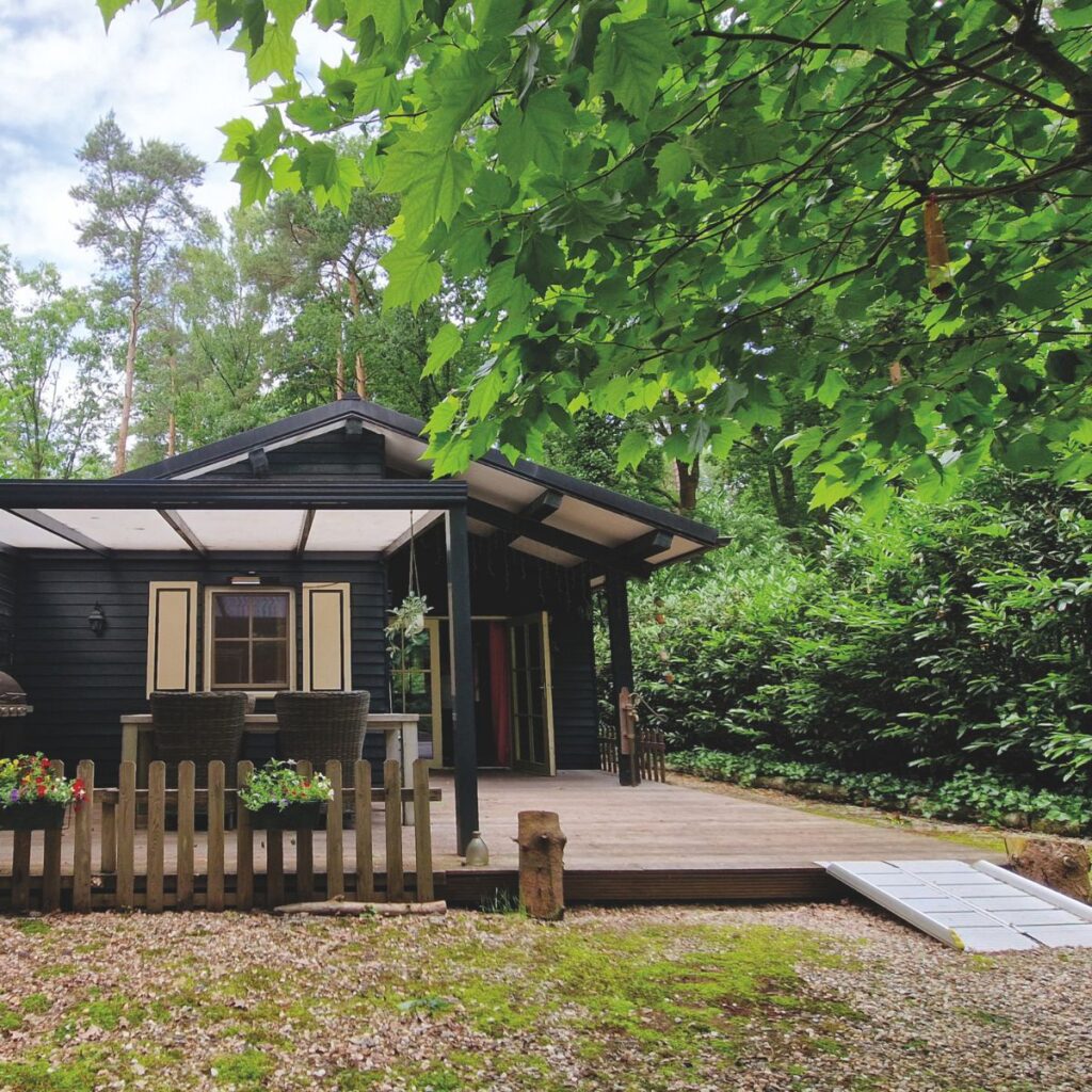 A side view of the cottage showing a wide wooden veranda. A metal threshold ramp connects the gravel driveway to the deck, providing a smooth transition for wheelchairs or walkers. A small wooden fence with flower boxes borders the seating area.