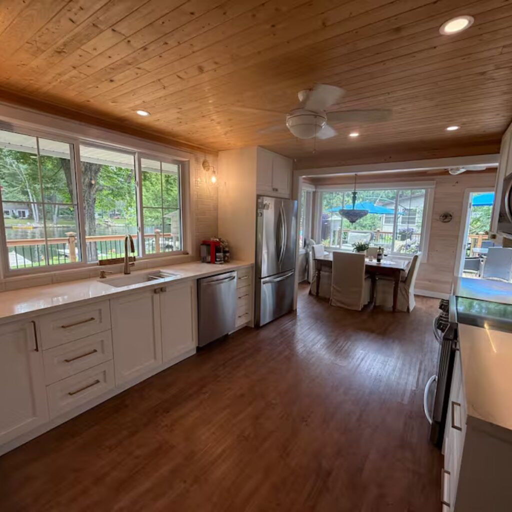 An open-concept kitchen and dining area with wood-look flooring and a wood-slat ceiling. The kitchen features white cabinetry and wide walkways. In the background, a dining table is set in front of large windows overlooking a lake, providing ample clearance for mobility.