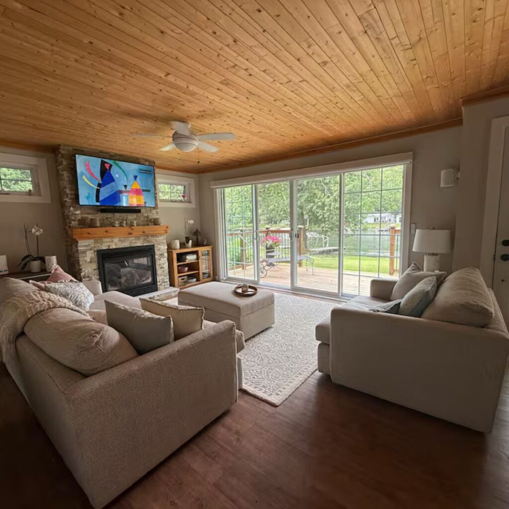 A cozy living room with a stone fireplace and a large TV mounted above it. Two neutral-toned sofas face each other over a patterned rug. Large sliding glass doors offer a level transition to an outdoor wooden deck with a scenic view of a river or lake.