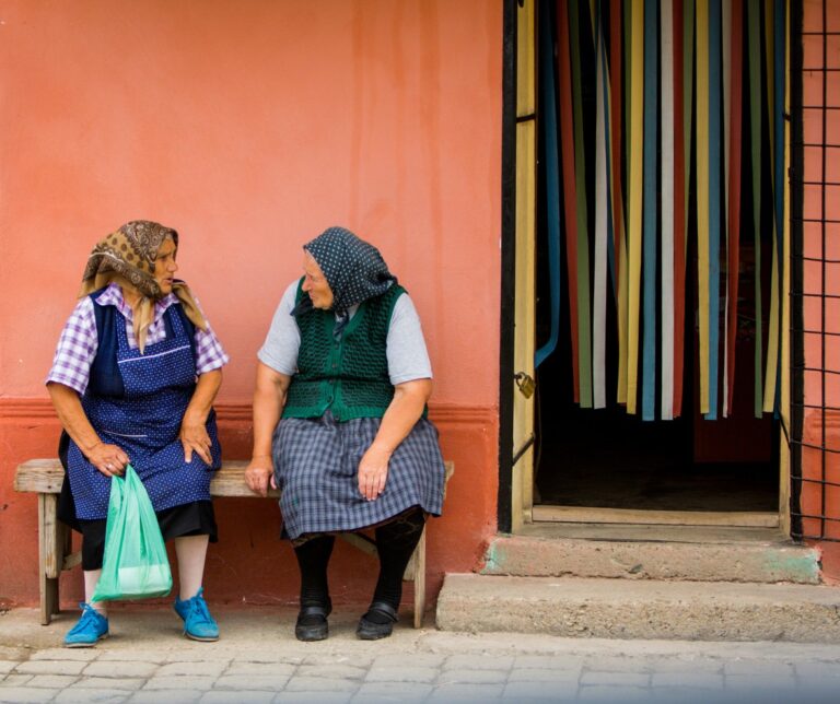Two elderly Romanian women in traditional headscarves sitting on a bench outside a colorful village shop in rural Transylvania, Romania