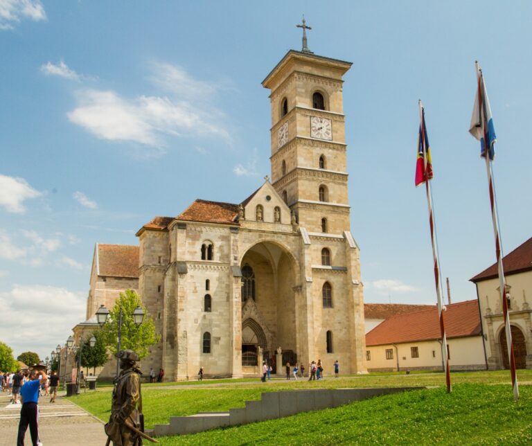 Romanesque exterior of the Roman Catholic Cathedral of Saint Michael in Alba-Iulia, Romania, with flags and a bronze statue in the foreground